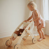 Child playing with a wooden doll stroller on a wooden floor.